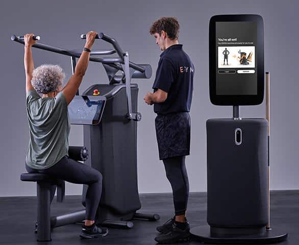 Oudere vrouw doet oefening op fitnessapparaat met personal trainer en scherm met instructies.