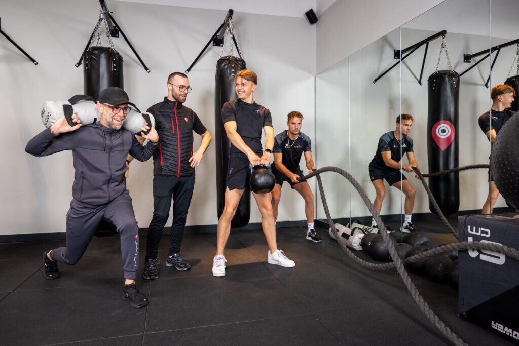 Vier mannen trainen in sportschool met bokszakken, zandzak, kettlebell en touw.