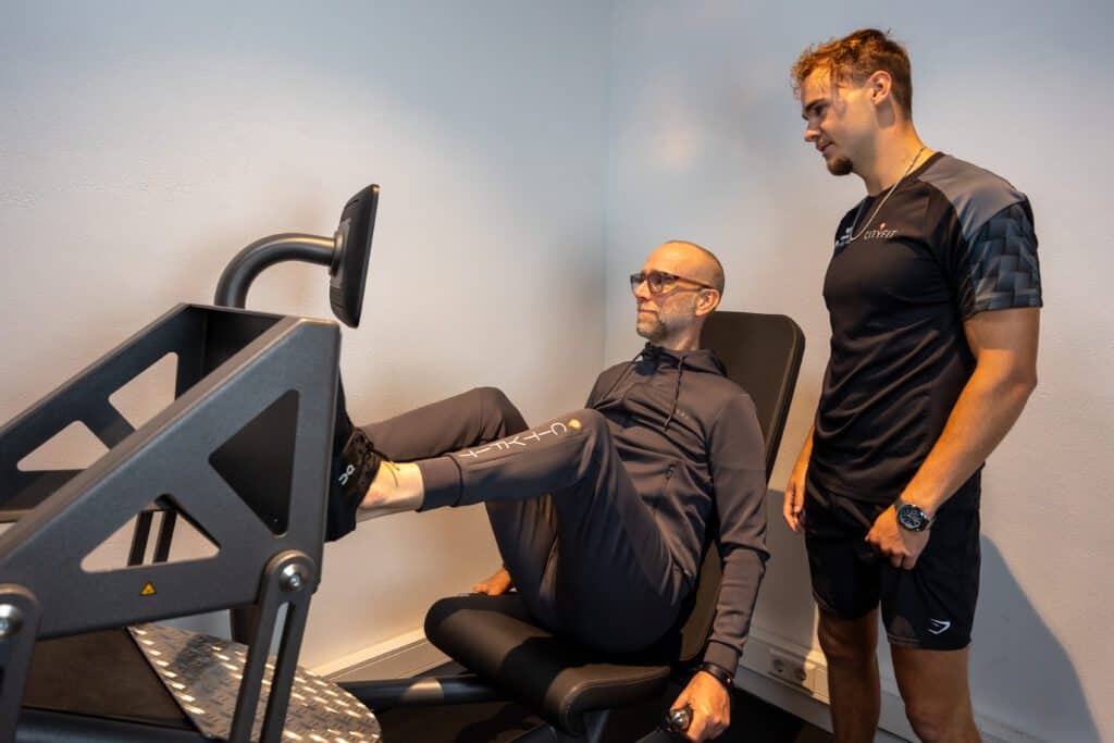 Man doet beenoefening op fitnessapparaat terwijl trainer toekijkt in sportschool.