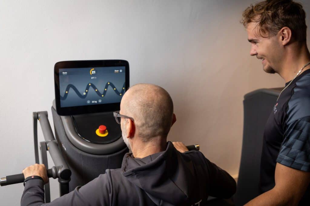 Oudere man gebruikt een EGYM apparaat, trainer begeleidt hem.